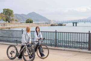 Dos personas en bicicleta junto a una orilla con un puente al fondo.