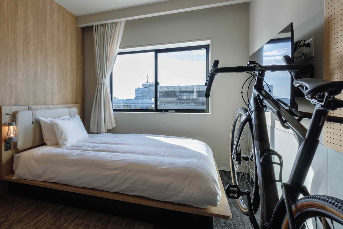 Une chambre d’hôtel avec un lit, un vélo, une fenêtre donnant sur la ville et une lampe fixée au mur.