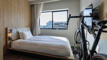 Une chambre d’hôtel avec un lit, un vélo, une fenêtre donnant sur la ville et une lampe fixée au mur.