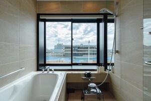 Ein modernes Badezimmer mit einer großen Badewanne, einer Dusche und einem Fenster mit Blick auf ein Kreuzfahrtschiff und Hafeneinrichtungen.