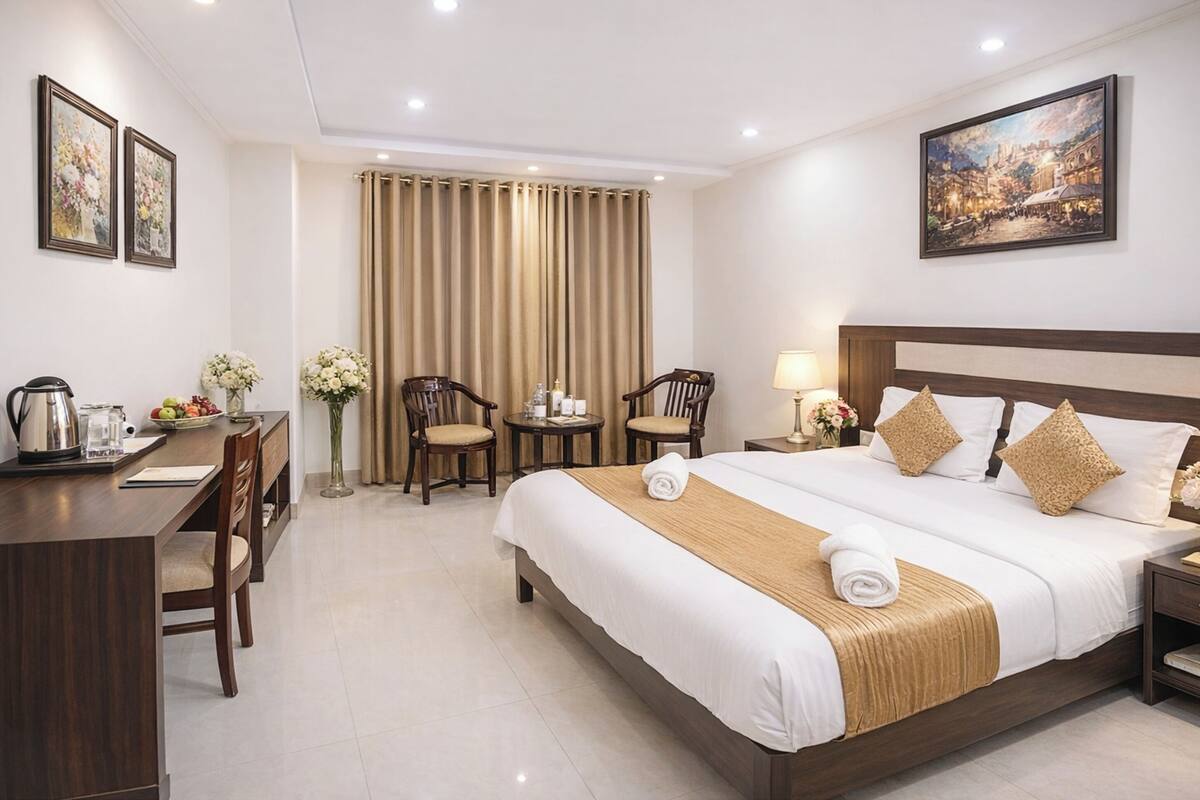 Quarto de hotel com uma cama grande, uma escrivaninha, duas cadeiras, uma mesinha, uma mesa de cabeceira e um quadro na parede.