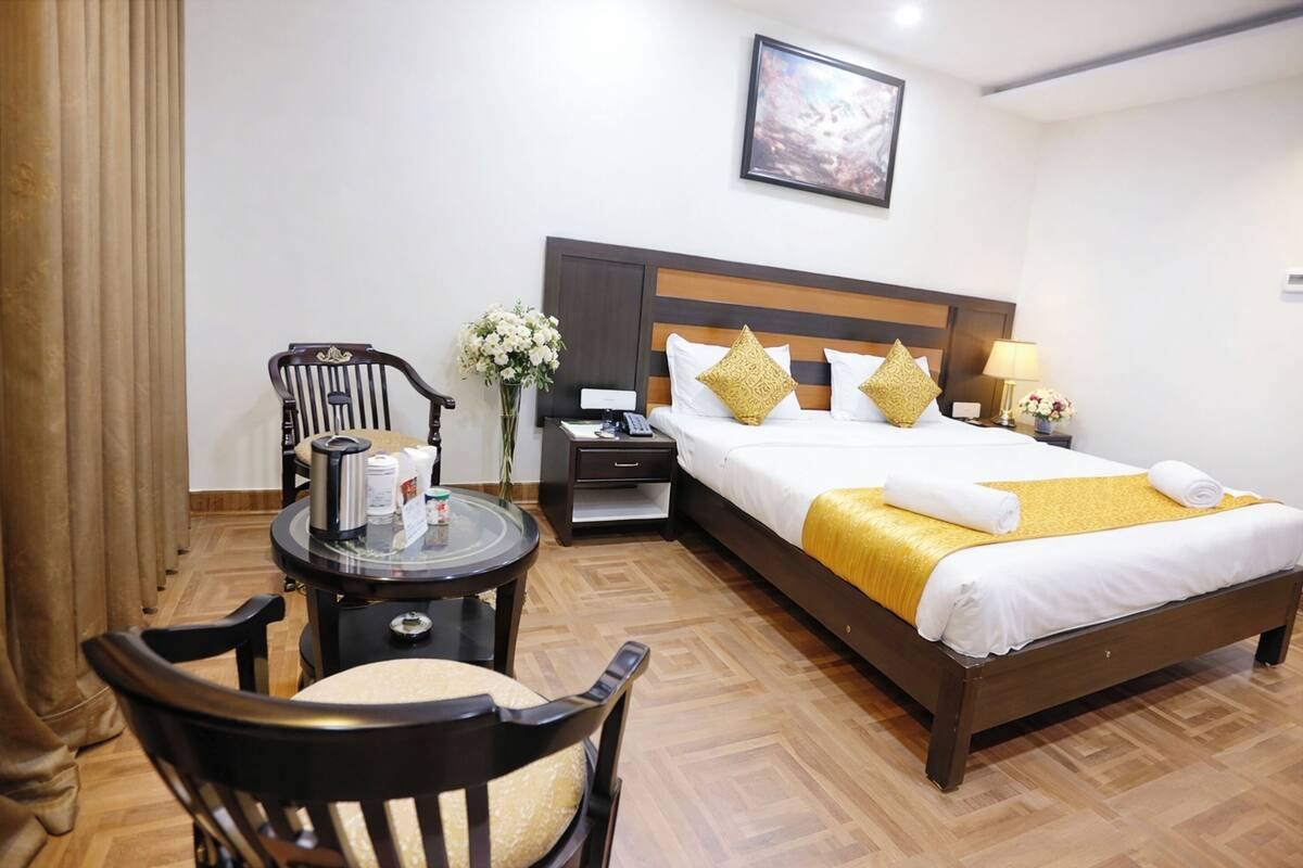 Quarto de hotel com cama de madeira, mesa de cabeceira com luminária, cadeira, pequena mesa com cafeteira e um vaso de flores.