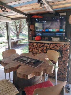 A wooden table with a flat-screen TV displaying a movie, a rustic chair, and a stack of firewood. - JAMIESON RIVERFRONT PROPERTY WITH FANTASIC OUTDOOR ENTERTAINMENT AREA (Jamieson)