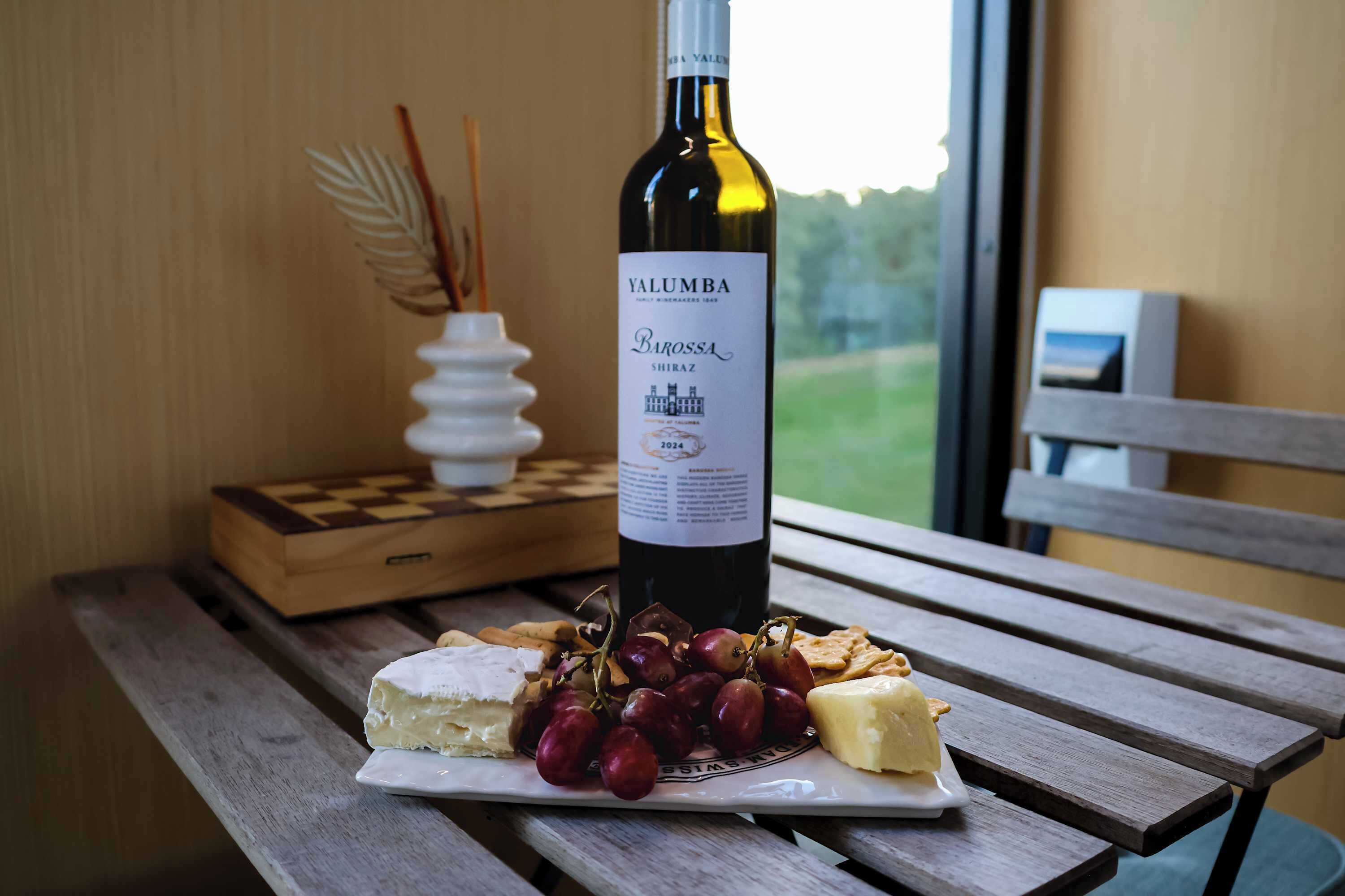 A wooden table with a bottle of wine, a plate with cheese and grapes, and a chessboard.
