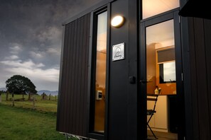 A small, dark wooden cabin with a lit interior, a sign indicating management, and a view of the outdoors.