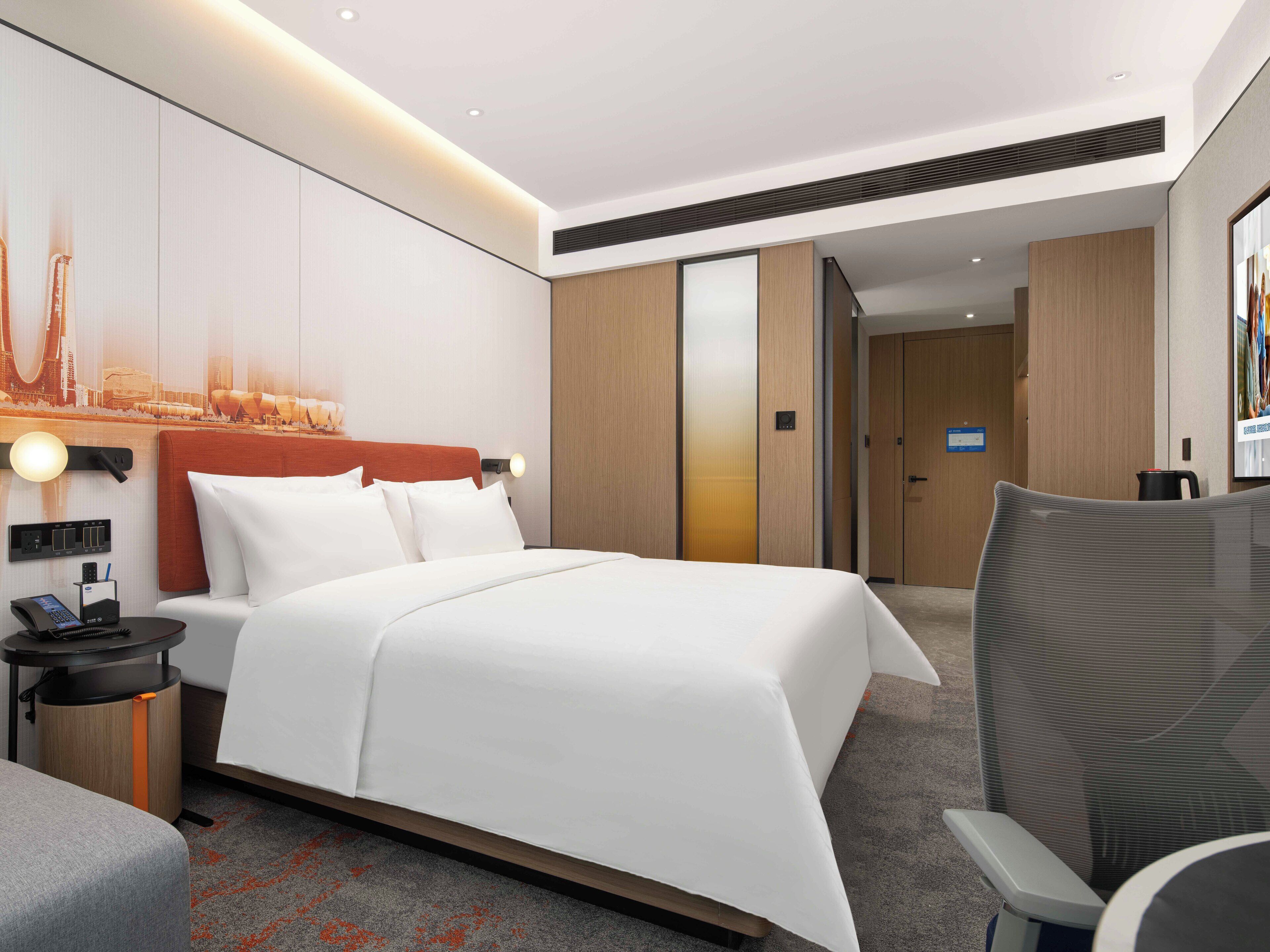 Ein modernes Hotelzimmer mit einem großen Bett, einem Schreibtisch mit Telefon, einem Sessel und einem großen Fenster.