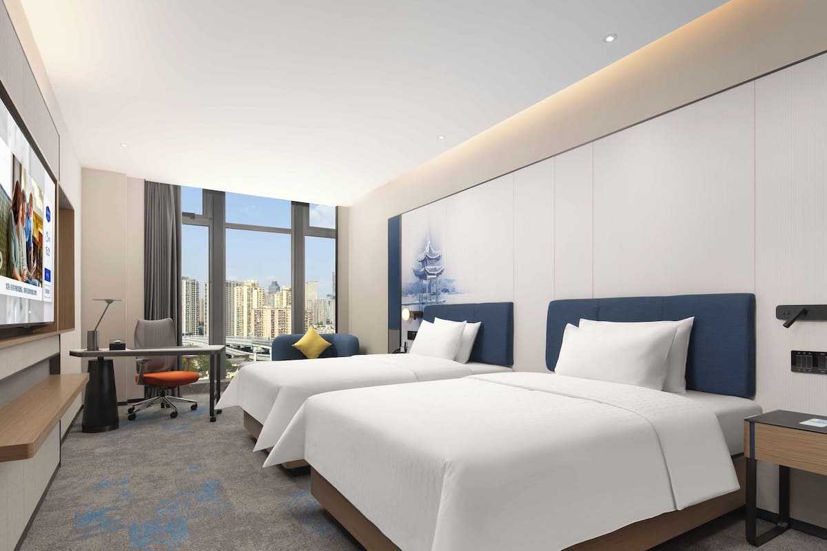 A hotel room with two beds, a desk, a chair, and a large window offering a city view.