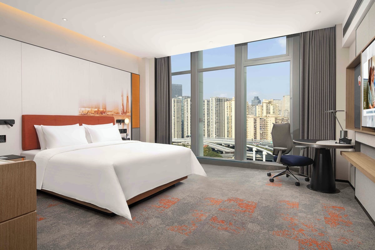 A hotel room with a bed, desk, and large windows offering a city view.