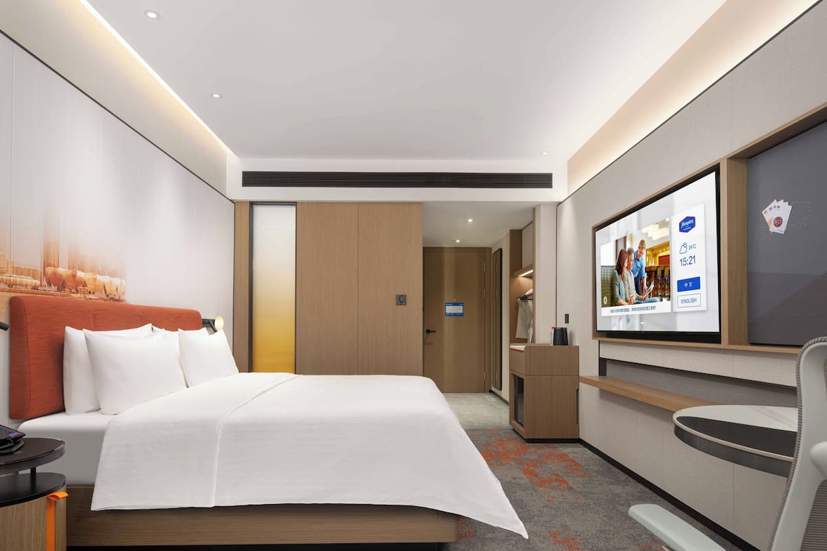 A modern hotel room with a large bed, a desk, a chair, a TV, and a view of the city through the window.