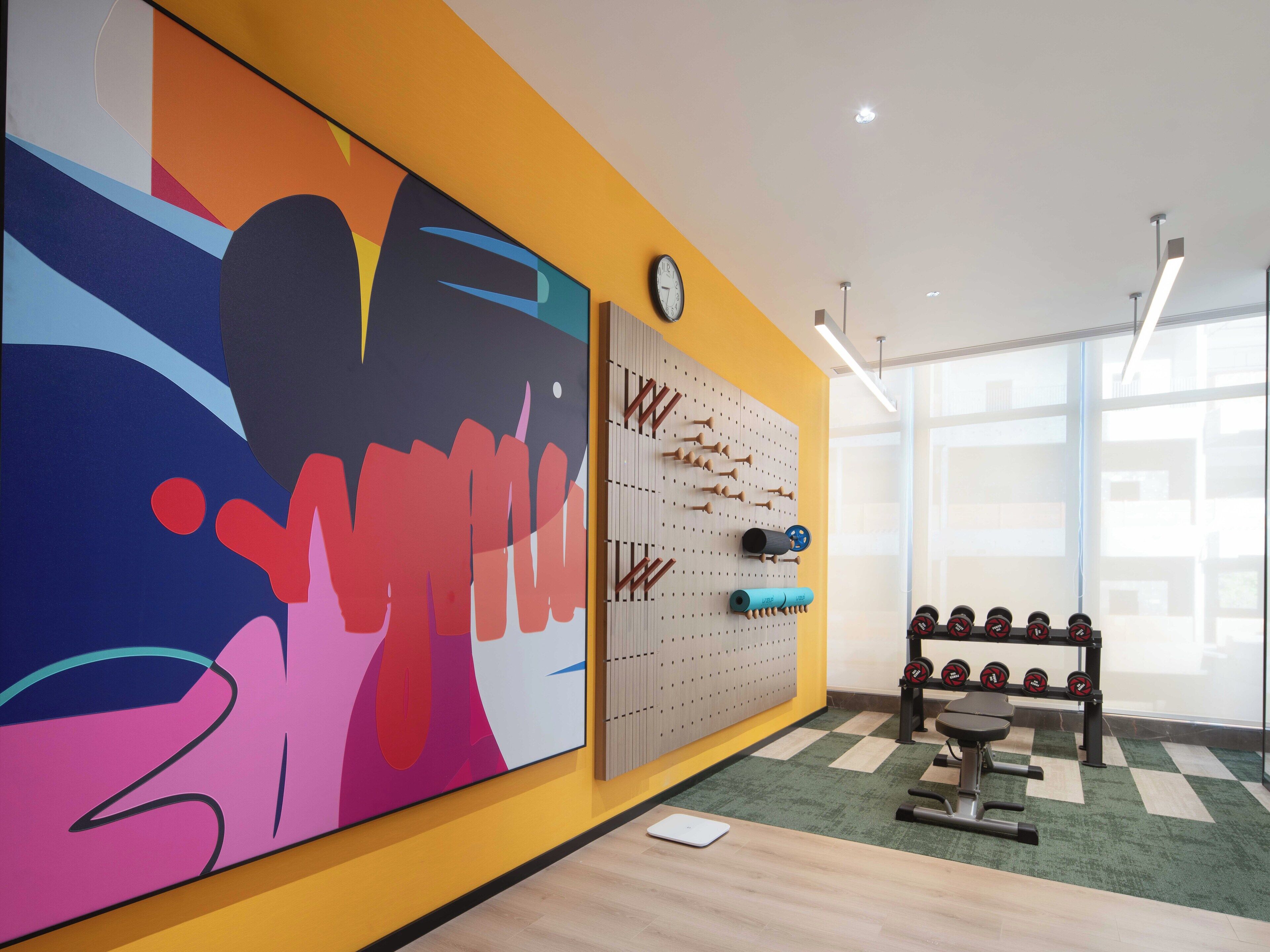 A gym with a colorful wall mural, a wall-mounted dumbbell rack, and a scale on the floor.
