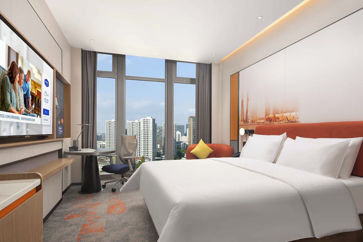A hotel room with a large bed, a desk, a chair, a TV, and a view of the cityscape.