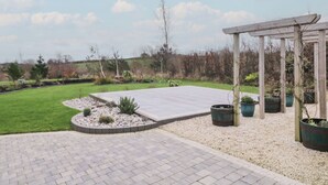A paved patio area with a pergola structure, surrounded by gravel and small plants. - The Loft - 96B (Coleraine)