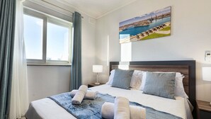 Een hotelkamer met een groot bed, een schilderij van een kustlandschap en een raam met gordijnen.