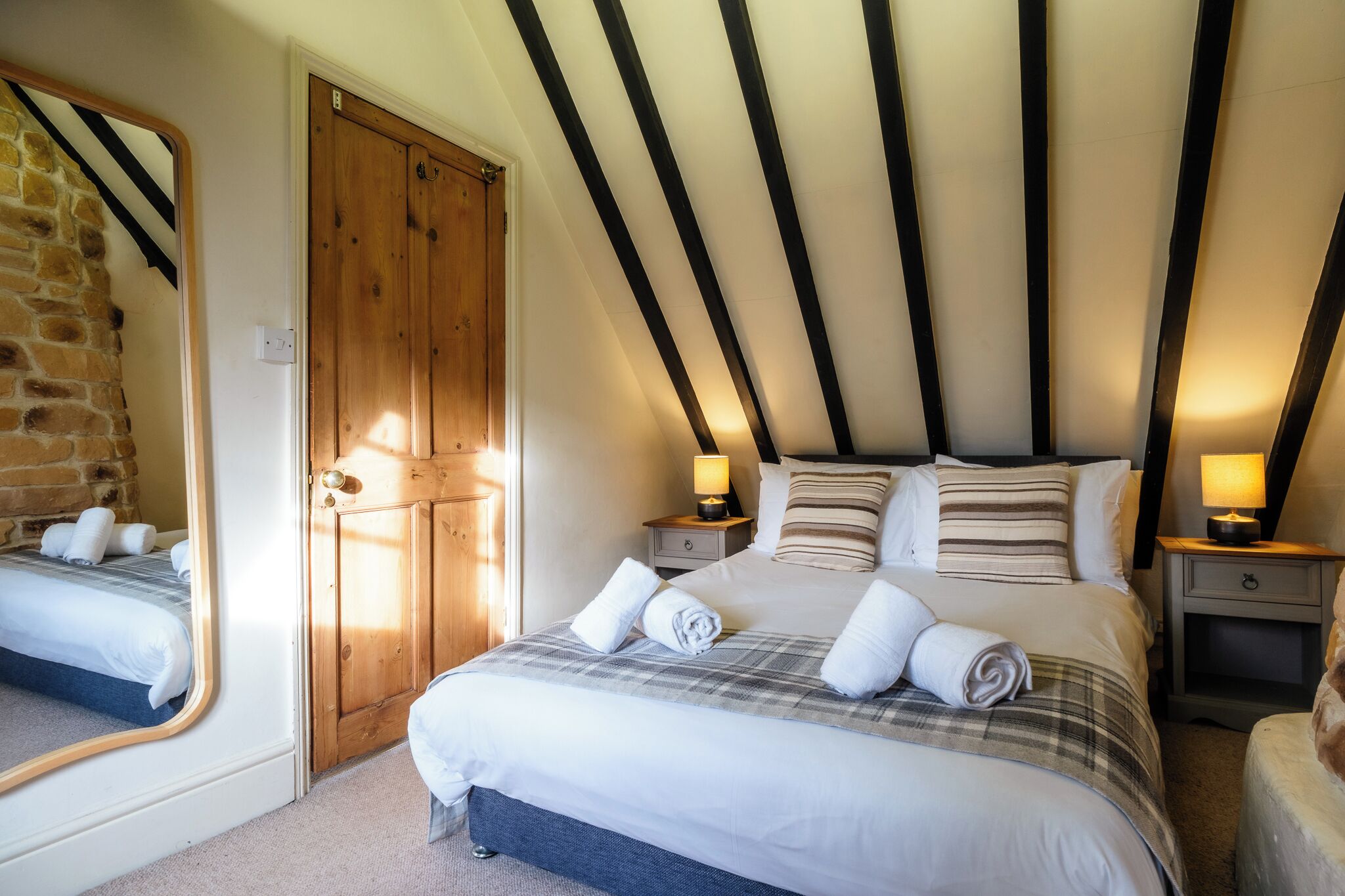 Een slaapkamer met een groot bed, houten balken in het plafond, een spiegel en twee nachtkastjes met lampen.