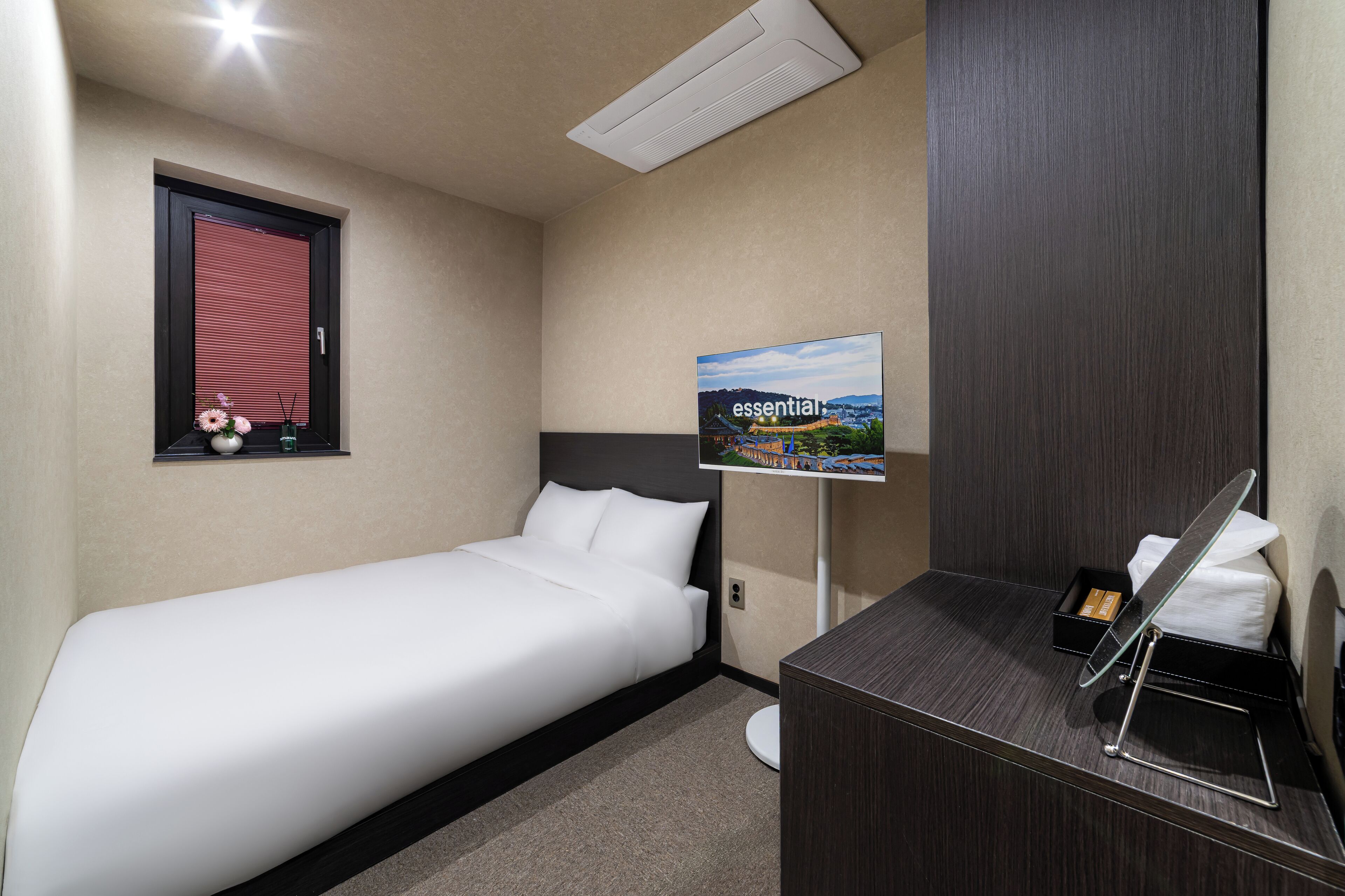 Een hotelkamer met een enkel bed, een klein bureau met spiegel, een wandairconditioner, een inlijsting foto en een raam met zonwering.