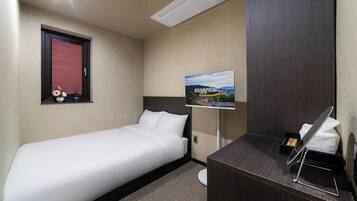 Een hotelkamer met een enkel bed, een klein bureau met spiegel, een wandairconditioner, een inlijsting foto en een raam met zonwering.