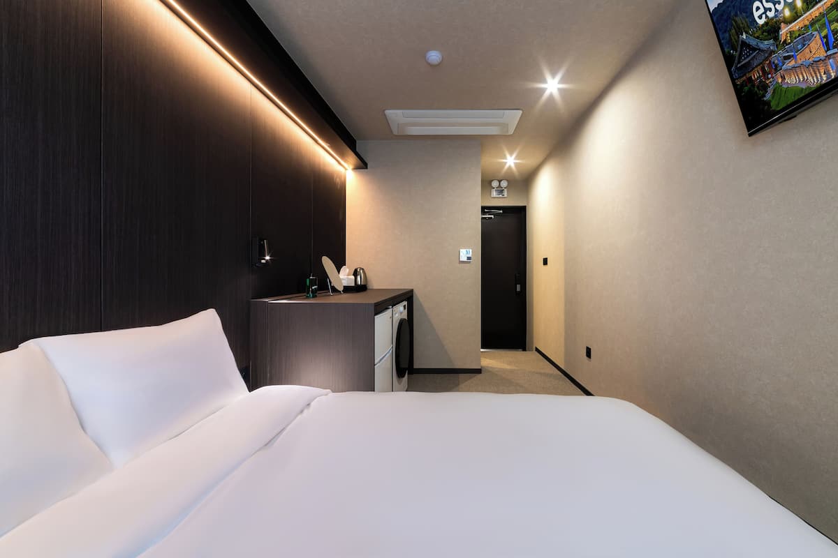Ein modernes Hotelzimmer mit einem ordentlich bezogenen Bett, einem kleinen Schreibtisch mit Telefon, einer Waschmaschine und einem an der Wand befestigten Fernseher.