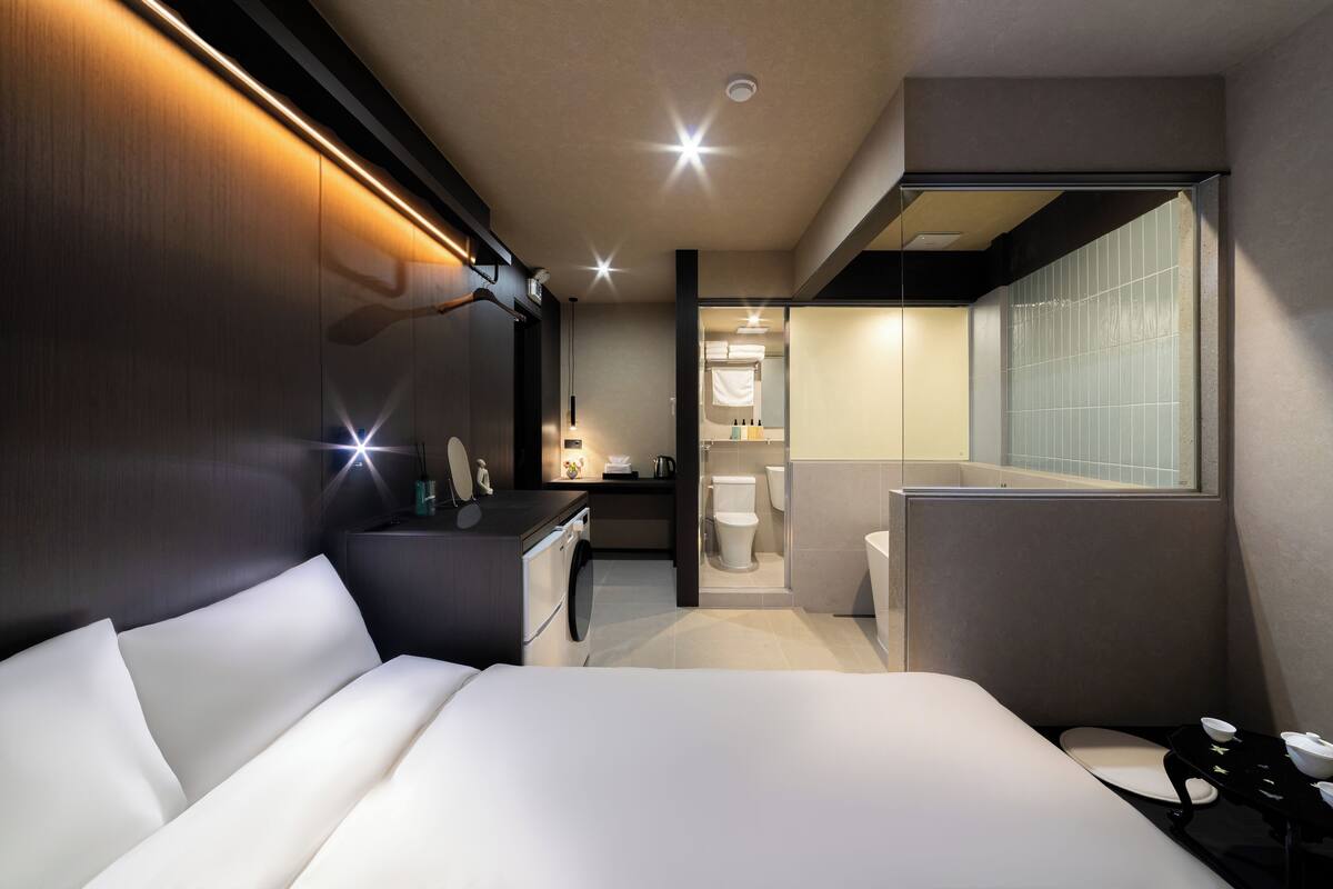 Ein modernes Hotelzimmer mit einem großen Bett, einem Schreibtisch, einem Spiegel und einem durch eine geöffnete Tür einsehbarer Badezimmer.