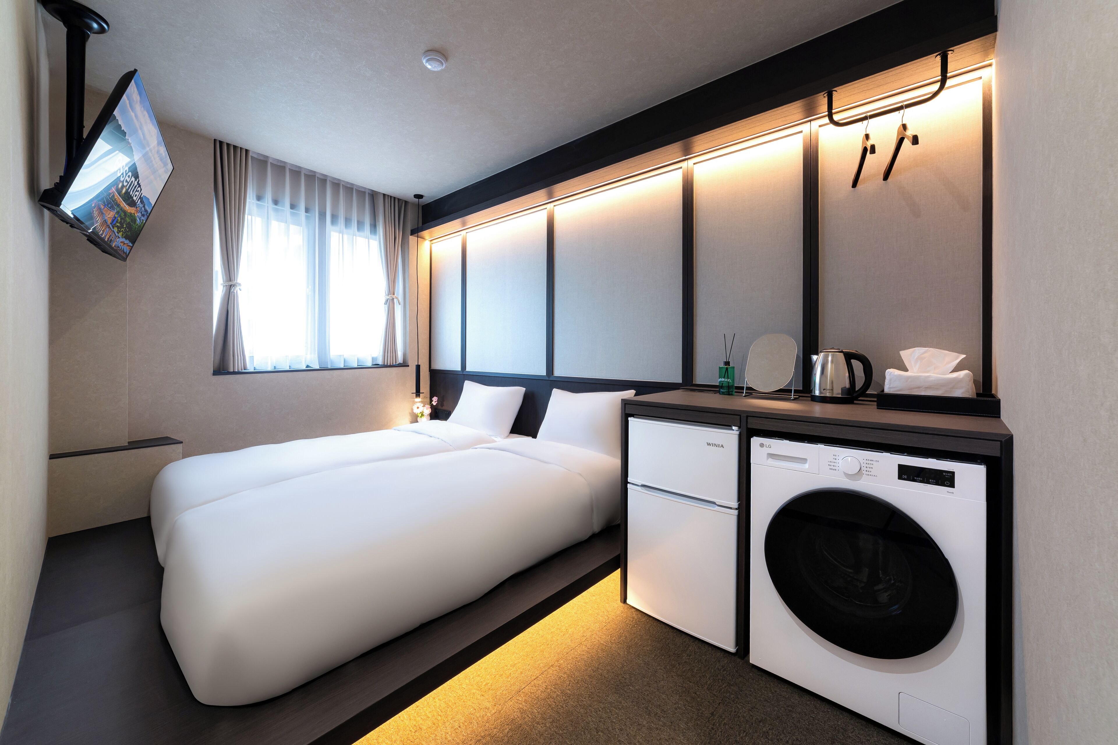 Habitación de hotel con cama, lavadora, una mesita con artículos de aseo y televisor montado en la pared.