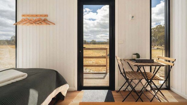 Un dormitorio con cama, una silla de madera y una mesa con una planta. Se ve una ventana con vistas al exterior.