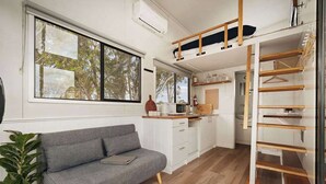 Un espacio habitable compacto con sofá, kitchenette y un altillo accesible mediante una escalera de madera.