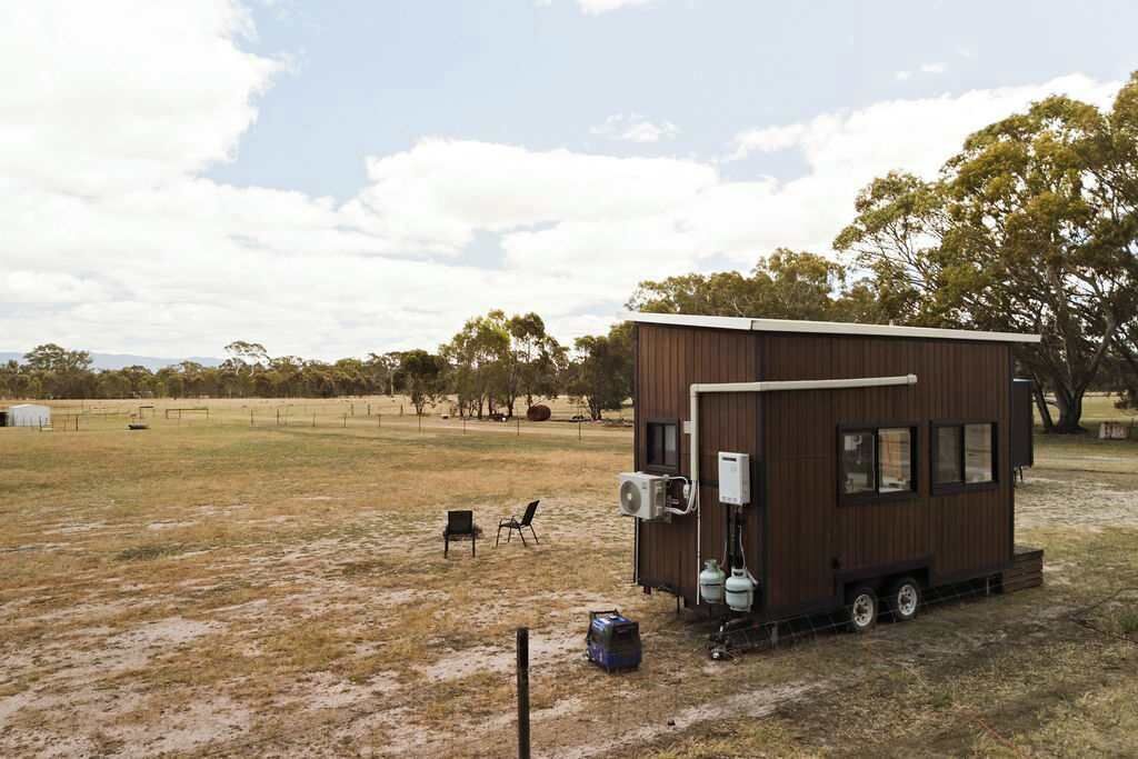 Une petite cabane en bois d’un seul étage, surmontée d’un toit en métal, située dans un champ herbeux avec des arbres en arrière-plan.