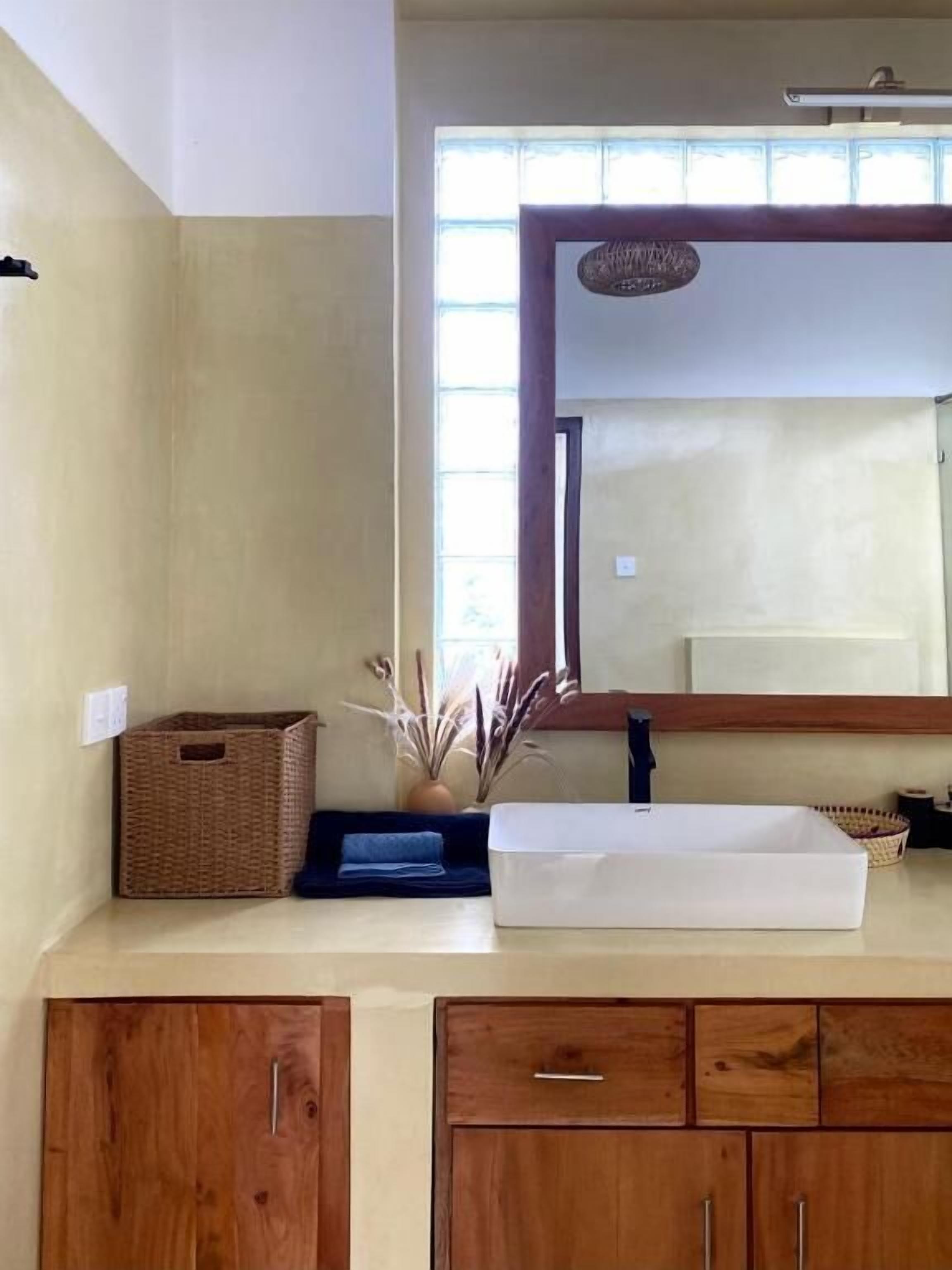 A bathroom with a large mirror, wooden cabinets, a wicker basket, and a white basin.