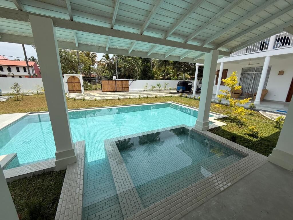 A swimming pool with a tiled deck, a covered patio area, and a view of a residential street.