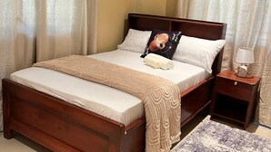 Un lit bien fait, avec une tête de lit en bois, une table de chevet avec une lampe et un tapis à motifs sur un sol carrelé.