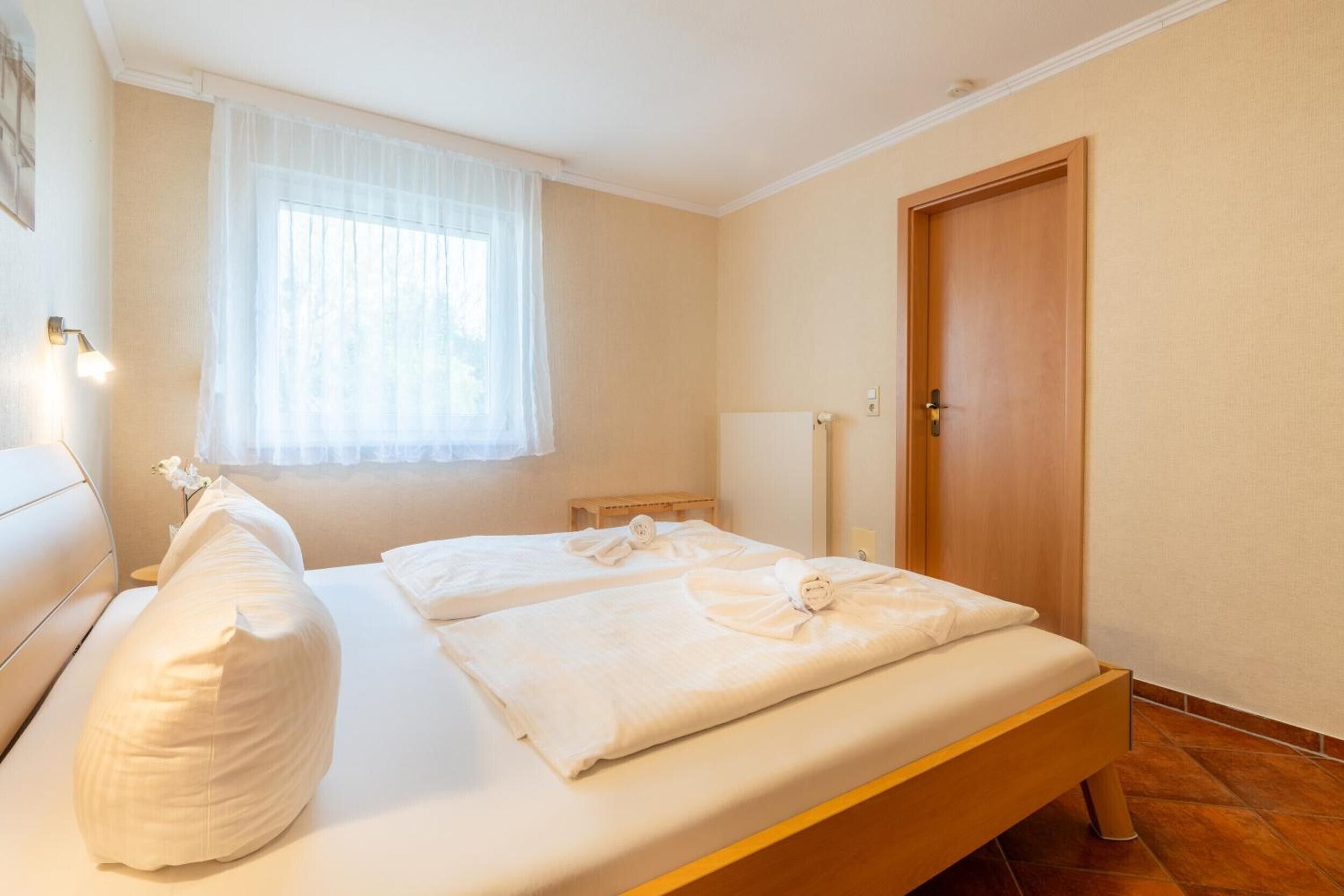 Ein Schlafzimmer mit zwei Betten, einer Holztür, einem Nachttisch und einem Fenster mit durchsichtigen Vorhängen.