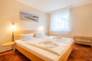 Quarto de hotel com cama, mesas de cabeceira, janela com cortinas translúcidas, um quadro na parede e um banco junto à janela.