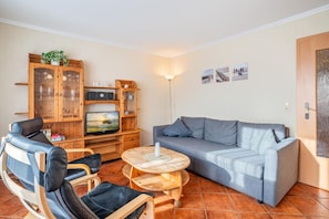 Uma sala de estar com sofá, duas cadeiras, televisão, armários de madeira e uma luminária.
