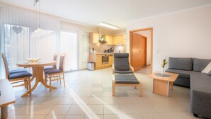 A modern living room with a sofa, a dining table with chairs, a kitchen area, and a large window with sheer curtains. - Cozy 2-Room Apartment on Usedom Island, Ground Floor, Perfect for 2 to 4 Guests (Pudagla)