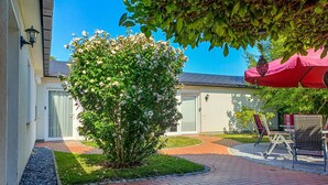 A courtyard with a brick pathway, a tree with white flowers, outdoor furniture, and a red umbrella. - Cozy 2-Room Apartment on Usedom Island, Ground Floor, Perfect for 2 to 4 Guests (Pudagla)