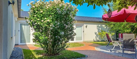 A courtyard with a brick pathway, a tree with white flowers, outdoor furniture, and a red umbrella.