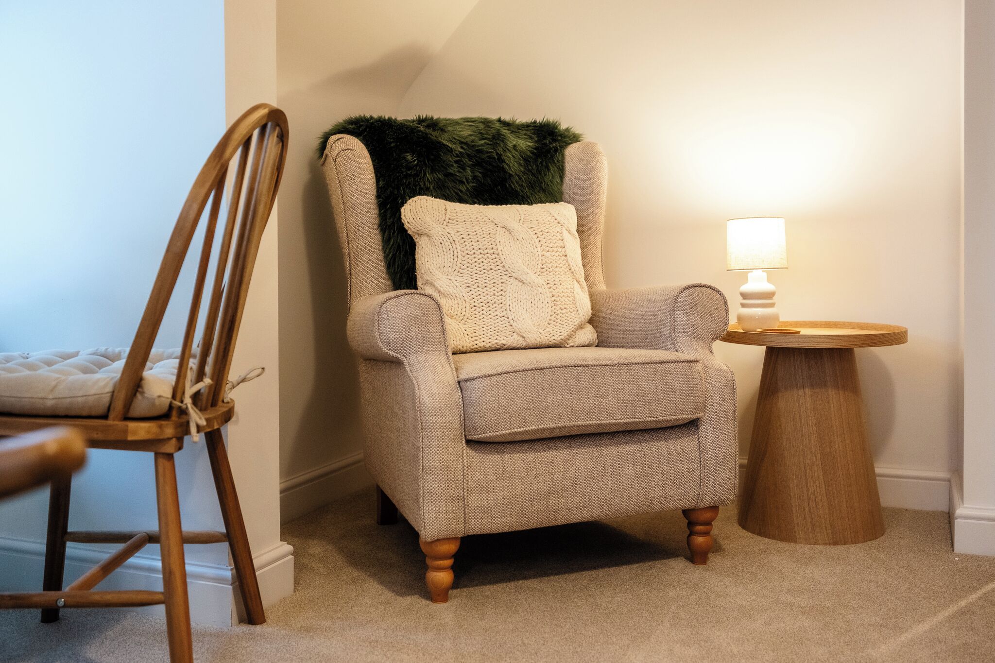 A corner room with a wooden chair, a cushioned armchair, a small wooden side table with a lamp, and a green furry throw on the armchair.