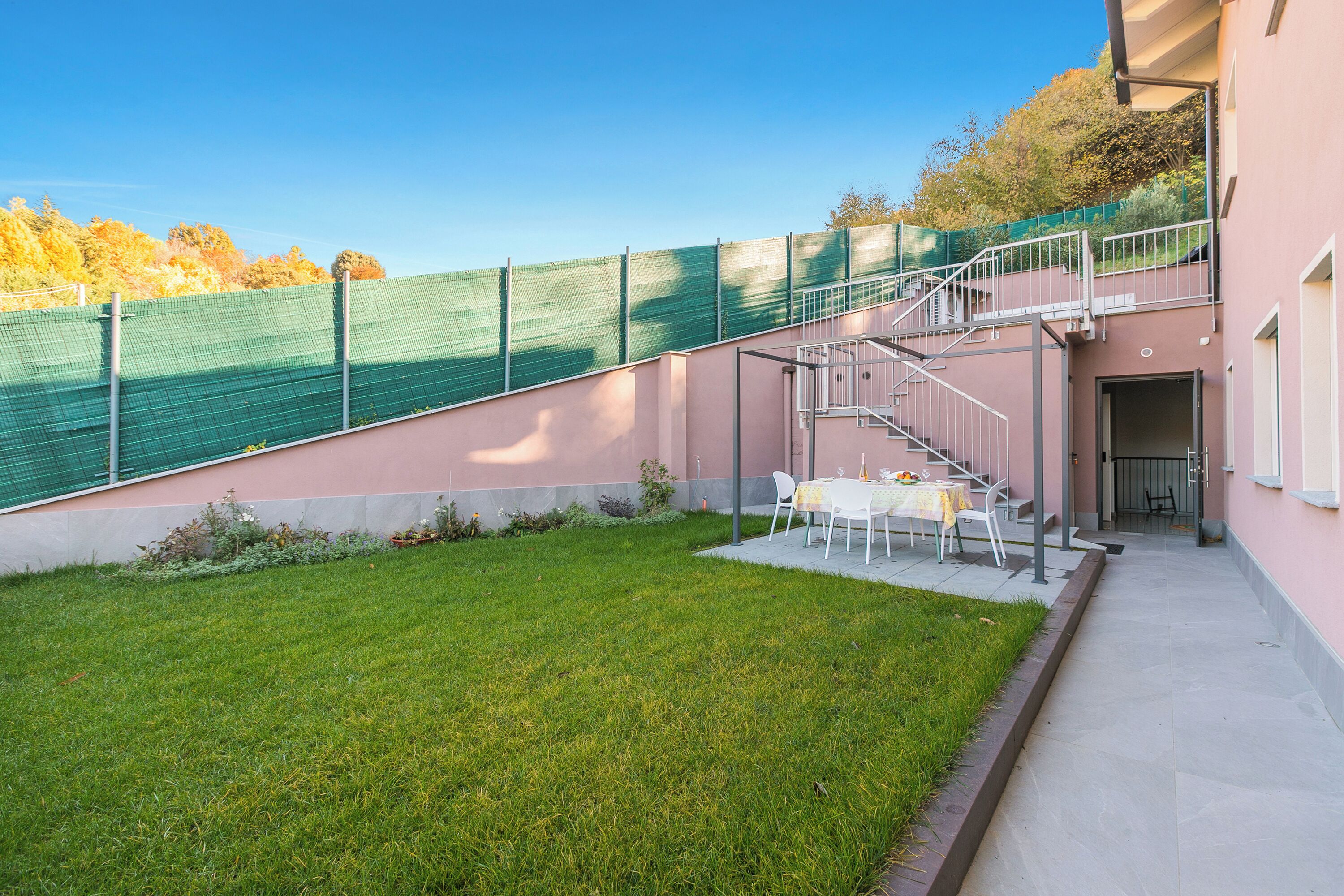 A building with a staircase leading to a balcony, a small outdoor seating area with tables and chairs, and a green fence.