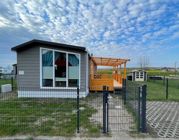 Une petite maison à un étage, dotée d’une cour clôturée, d’une terrasse en bois et surplombant un ciel dégagé.