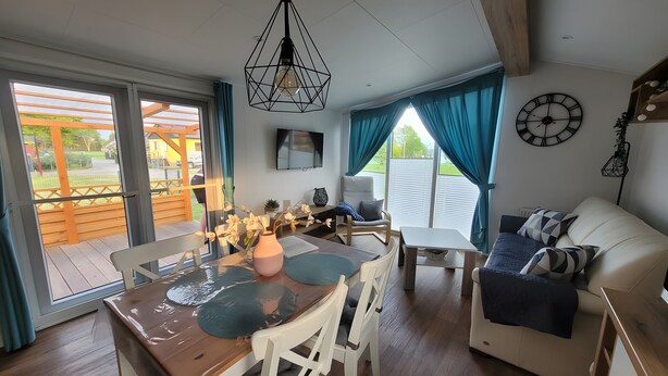 A modern living room with a dining area, wooden flooring, a ceiling light fixture, a TV, and a clock on the wall. - Holiday Home Concordia by the Lake with Covered Terrace and Air Conditioning (Seeland)