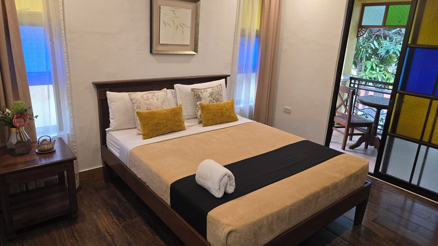 A neatly made bed with a black and beige bedspread, two pillows, and a folded towel. A small table with a vase and a teapot is beside the bed.