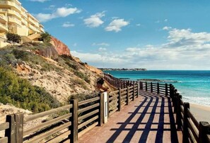 Ein hölzerner Steg führt zu einem Strand mit einer felsigen Hügelkuppe und einem Gebäude im Hintergrund.
