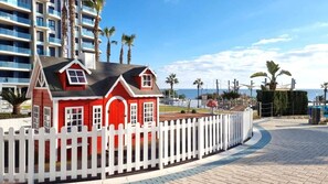 A red house with a white picket fence, surrounded by palm trees and a beach view. - Luxury seafront apartment La Zenia – Sea Senses (Torrevieja)