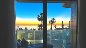 Ein Balkon mit freiem Blick auf den Sonnenuntergang über dem Meer, mit Palmen und einer entfernten Brücke.