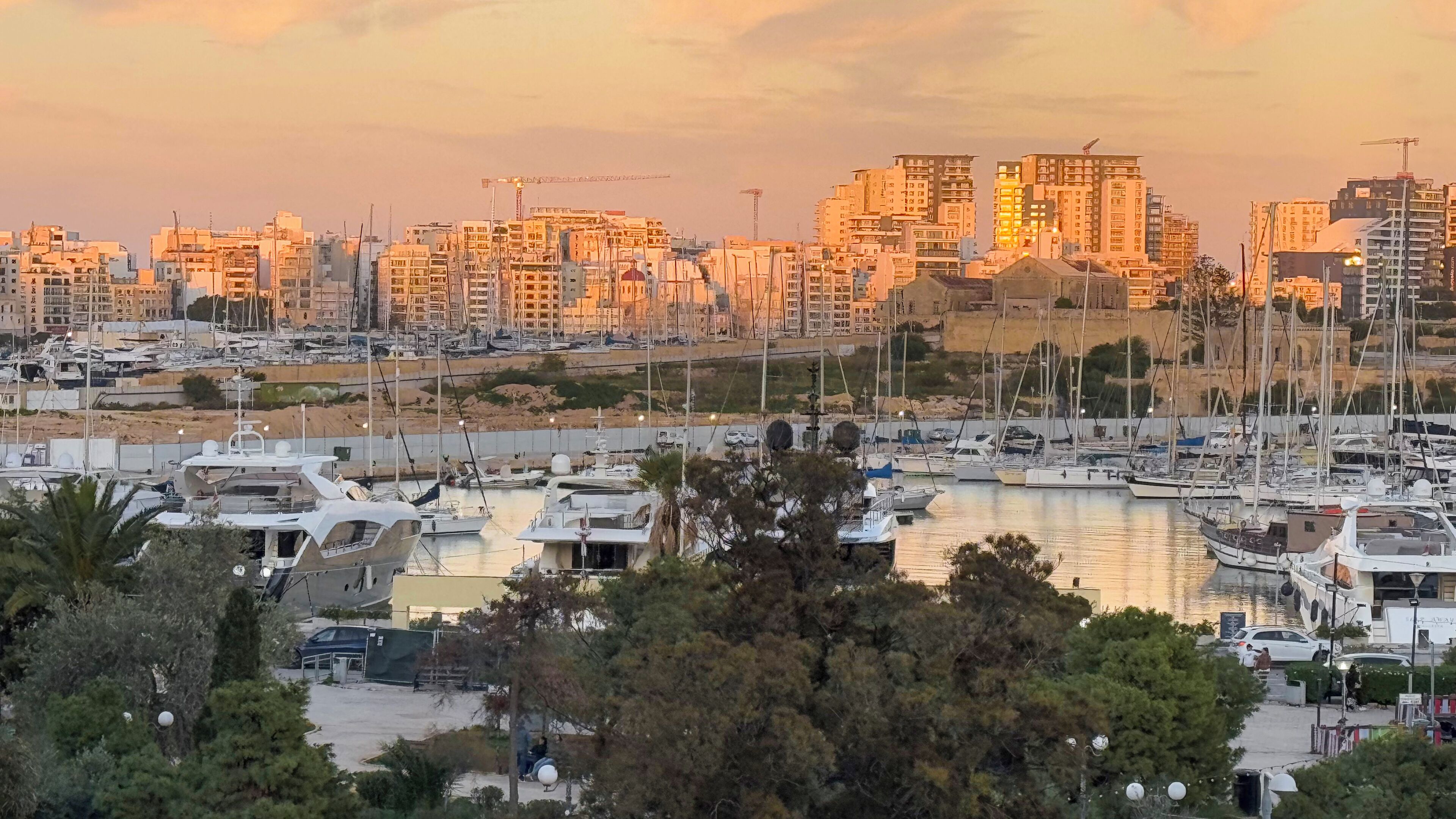 Un puerto deportivo con barcos amarrados, rodeado de edificios residenciales y grúas de construcción.