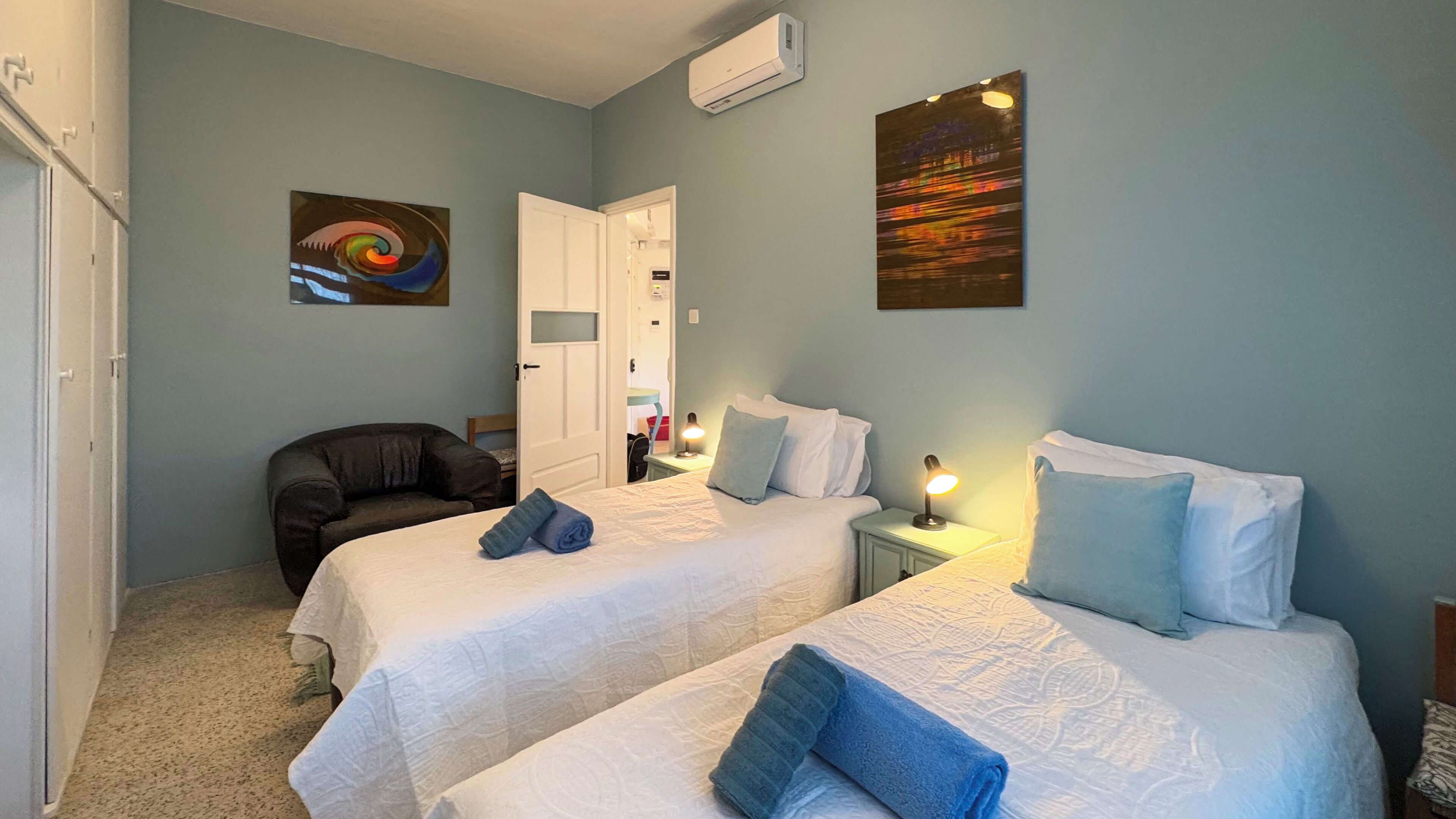 Une chambre d’hôtel avec deux lits, une chaise, un bureau et des œuvres d’art accrochées aux murs.