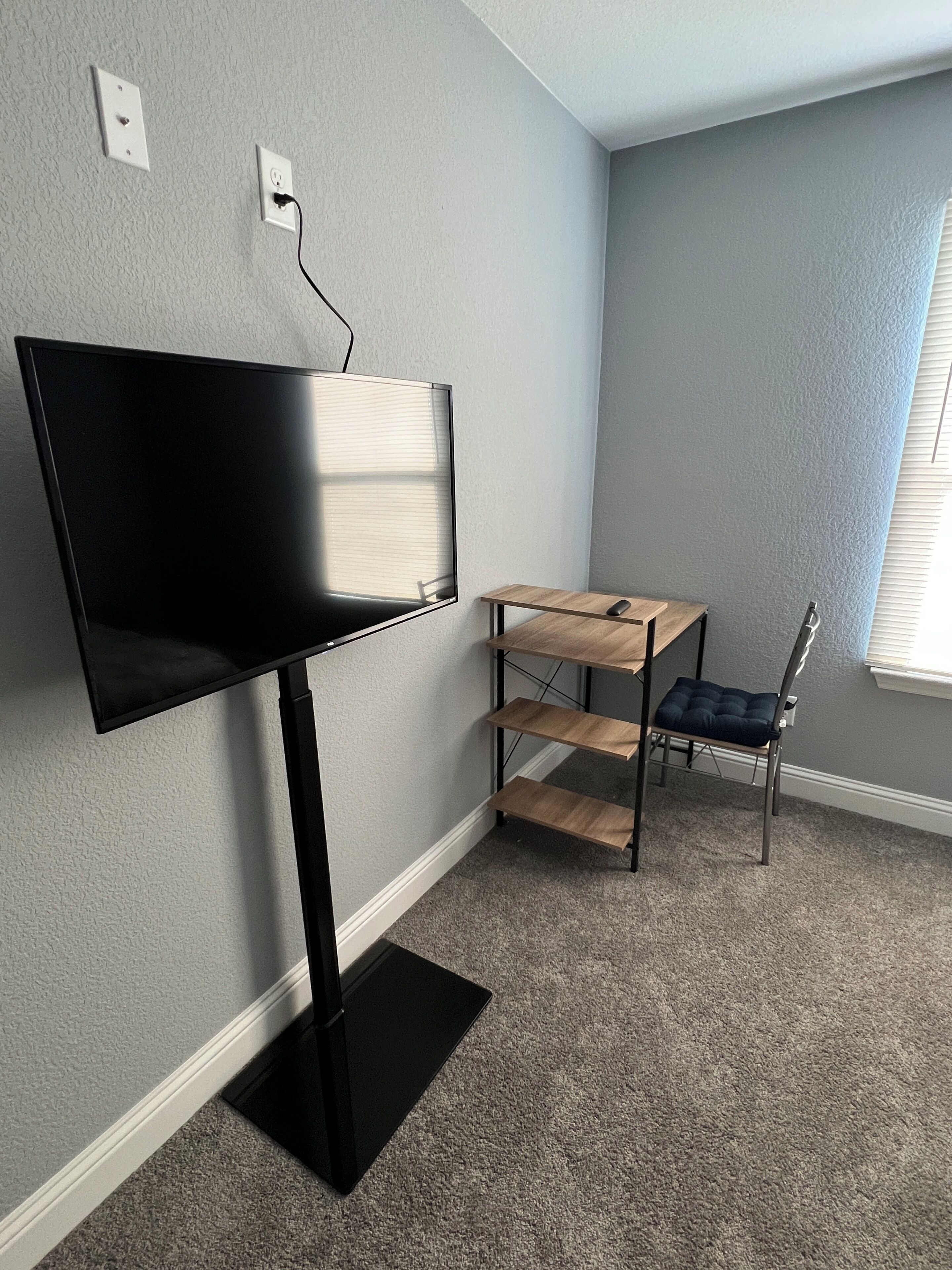 A room with a flat-screen TV mounted on the wall, a wooden desk with a chair, and a carpeted floor.