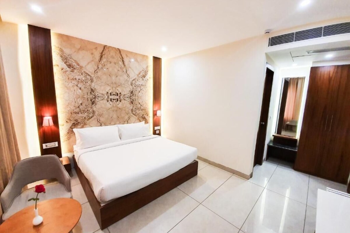 Habitación de hotel con una cama, una mesita con un florero, una silla, pared de mármol y puerta de madera.