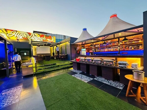 Zona de bar al aire libre con un letrero que dice 'High Lounge', una barra con varios artículos y una zona de estar con suelo estampado.