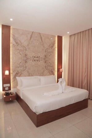 Una cama grande con estructura de madera, cabecera con estampado de mármol y colcha blanca. Hay dos mesitas de noche con lámparas, un aparador de madera y una cortina color beige.
