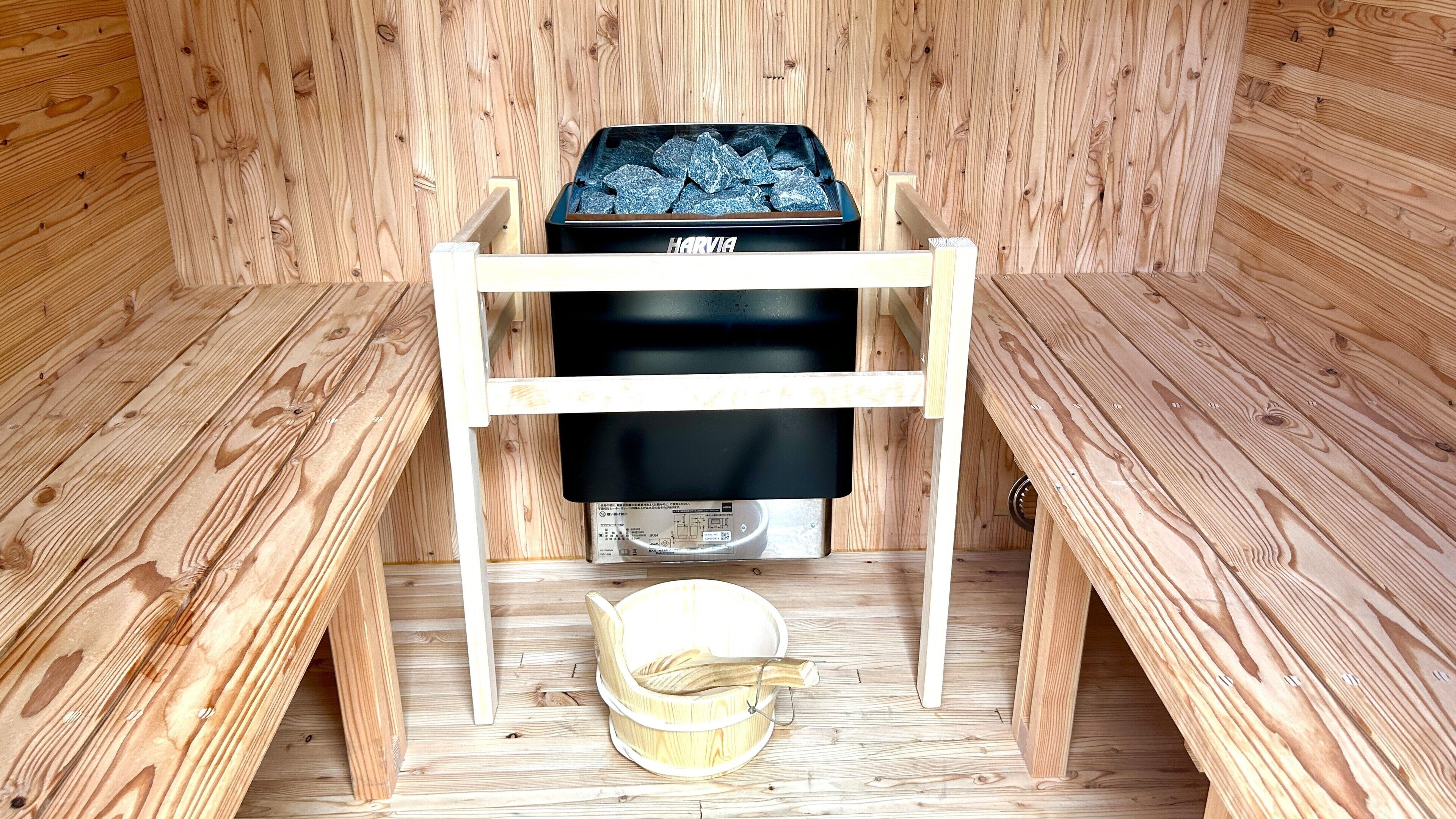 Een sauna met een zwarte heater, houten banken en een houten emmer.